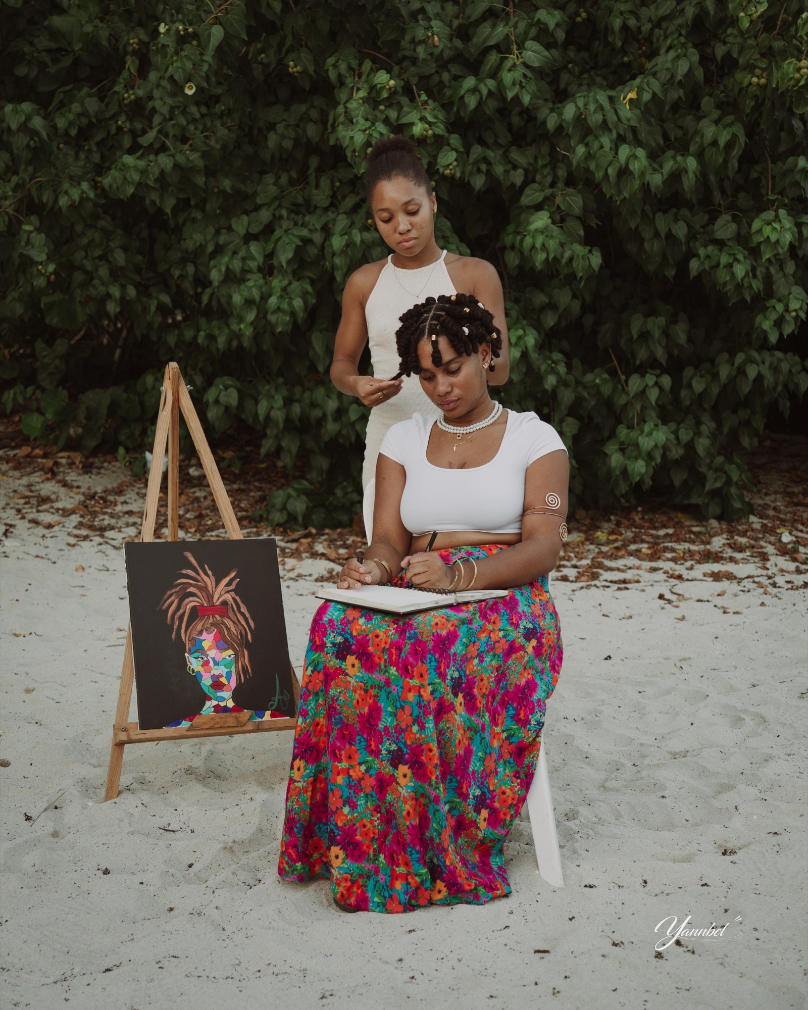 Aurianne dessine pendant qu’une coiffeuse réalise sa coiffure sur une plage de Guadeloupe